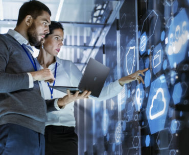 Male IT Specialist Holds Laptop and Discusses Work with Female Server Technician. They're Standing in Data Center, Rack Server Cabinet with Cloud Server Icon and visualization