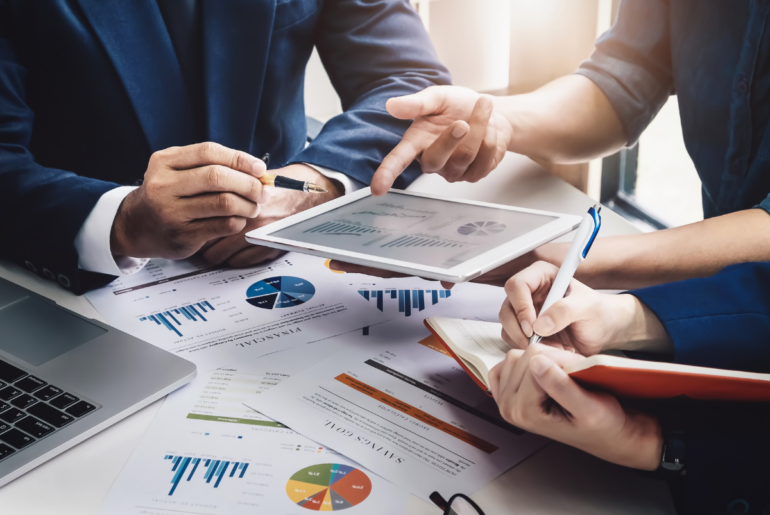 Business people, one in a suit, others dressed casually, engaged in discussion around a table full of data reports.