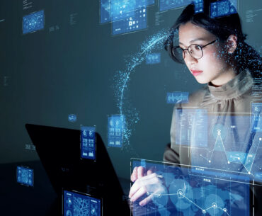 Woman with glasses looking at computer screen with holograms of data charts.