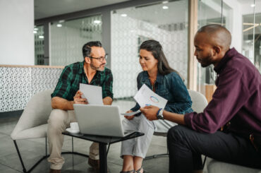 Three business professionals reviewing financial charts and data on a laptop during a strategy meeting.