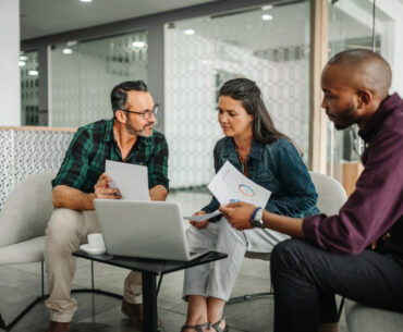 Three business professionals reviewing financial charts and data on a laptop during a strategy meeting.
